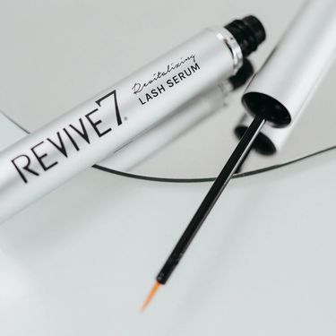 A silver tube of Beauty D Revive 7 Lash Revitalizing Serum 3mL with its applicator wand rests on a round mirror, emphasizing this lash serum formulated to support eyelash growth.